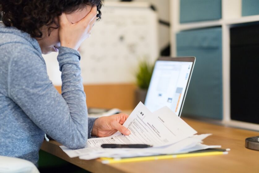 A young woman sits at a desk late at night and tries to work on her taxes. She looks discouraged as she reads a lt16 IRS notice and other paperwork. Her laptop is open in the background.