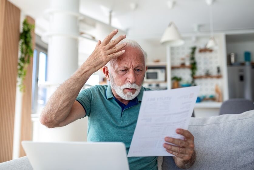 Worried senior man reading utility bill and using laptop at home. Stressed elderly male looking at paper with debt or high taxes. Financial problem, bankruptcy and money crisis concept for seniors.