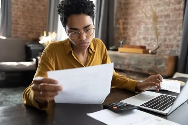 Woman stares intently at a document while sitting at her desk with a laptop and a calculator