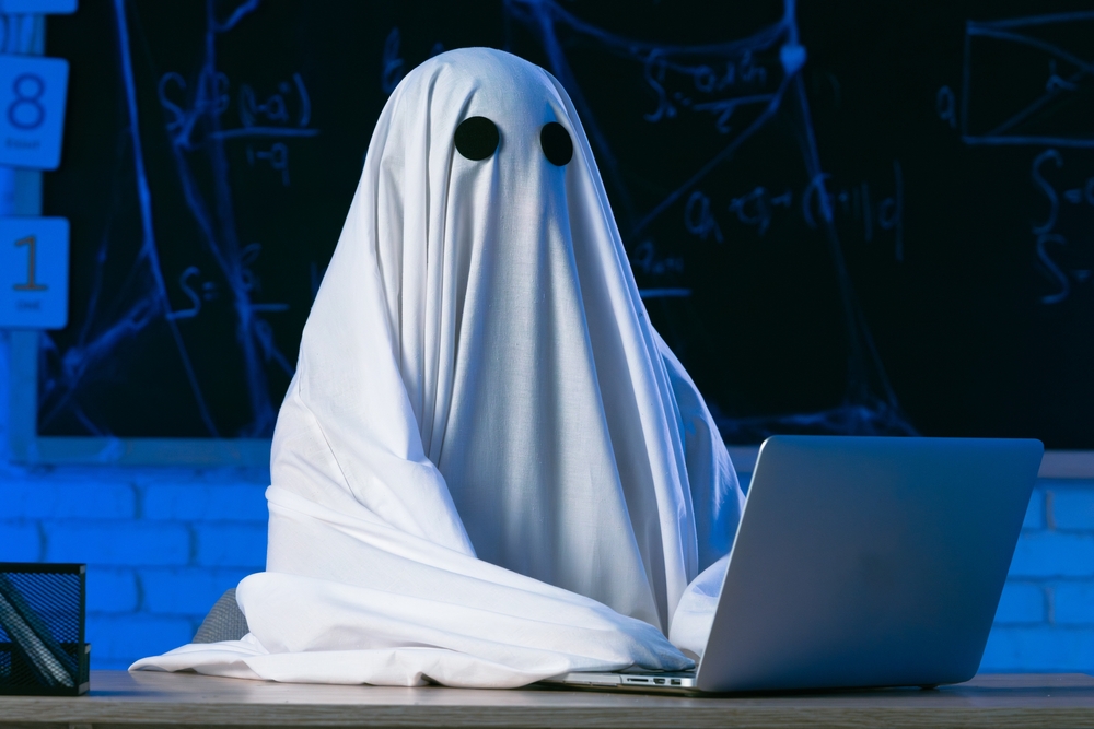 A person in a white ghost sheet sits at a table in a blue-lit room and types on a laptop