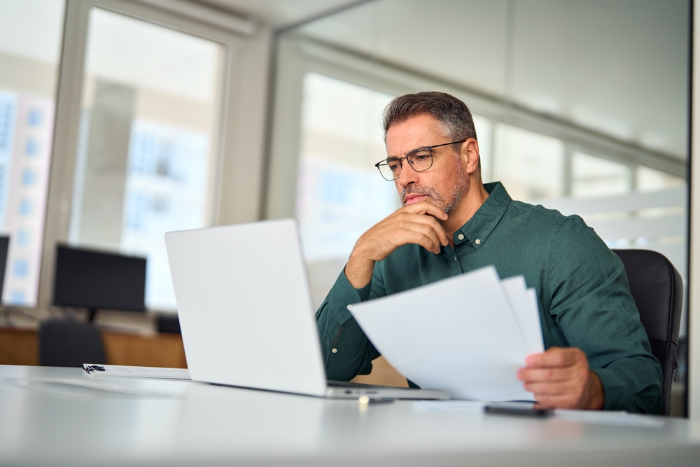 Busy middle aged executive, mature male hr manager holding documents using laptop looking at pc in office at desk, thinking over financial data report feeing doubt about market assets investment risk.
